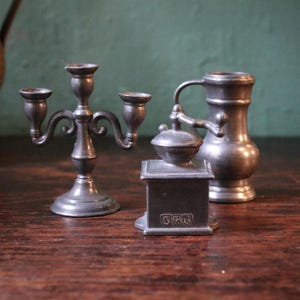 May include: A collection of antique pewter items, including a three-branch candelabra, a miniature coffee grinder, and a small jug. The objects are displayed on a dark wooden surface with a green background. The coffee grinder has a crank.