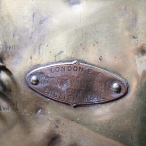 May include: Close-up of a brass object with a copper-coloured oval plaque. The plaque reads "LONDON E.C. ROTAX MOTOR CO. 508 ENGLISH MADE". The object has a tarnished appearance and visible rivets.