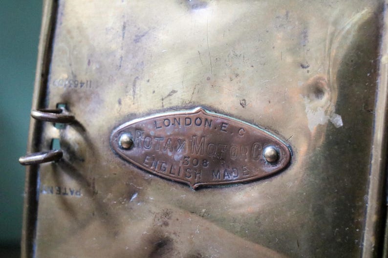 May include: Close-up of a brass object with a copper-coloured oval plaque. The plaque reads "LONDON E.C. ROTAX MOTOR CO. 508 ENGLISH MADE". The object has a tarnished appearance and visible rivets.