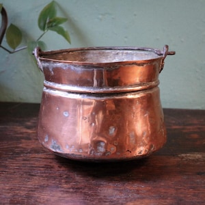 May include: An aged copper pot with a rounded base and two small handles. The pot shows signs of wear and a dark patina, with a lighter copper interior. The pot's exterior is a rich, reddish-brown colour, suggesting its age and use.