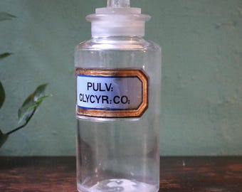 May include: A clear glass apothecary bottle with a glass stopper. The bottle has a blue and gold label with the text "PULV: CLYCYR:CO:". The bottle is cylindrical with a rounded base and is sitting on a wooden surface.