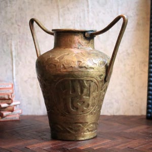 Puede incluir: Un jarrón de latón antiguo con dos asas arqueadas. El jarrón presenta intrincados diseños florales y geométricos en relieve. El metal envejecido tiene un tono dorado cálido, lo que sugiere que es un objeto decorativo antiguo.