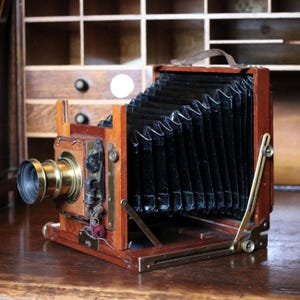 May include: A vintage wooden camera with a brass lens and bellows. The camera has a dark brown leather handle and is set against a wooden background. The camera's design is reminiscent of early photography equipment.