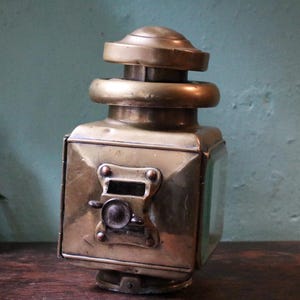 May include: Vintage brass lantern with a square body and a domed top. The lantern has a small, round handle and a glass panel. The brass has a warm, aged patina, suggesting its age. The lantern is a decorative item.
