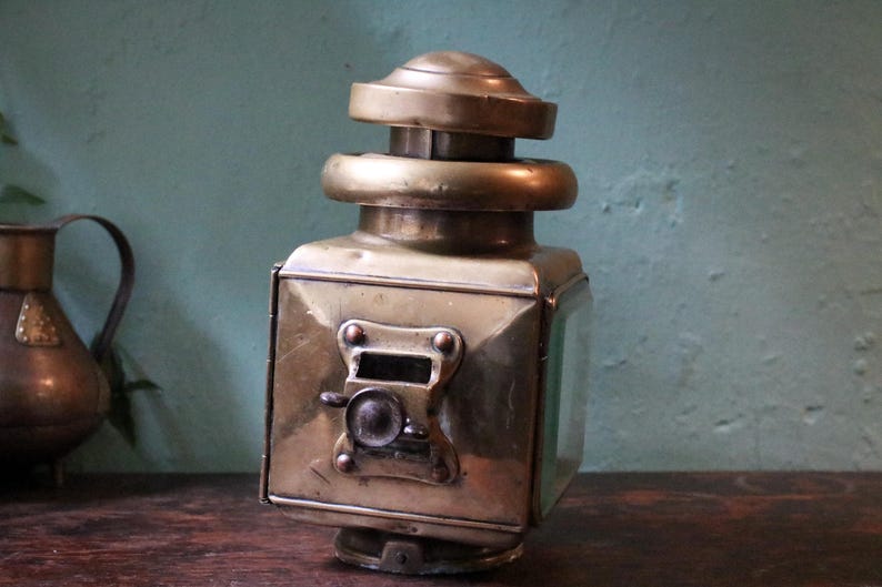 May include: Vintage brass lantern with a square body and a domed top. The lantern has a small, round handle and a glass panel. The brass has a warm, aged patina, suggesting its age. The lantern is a decorative item.