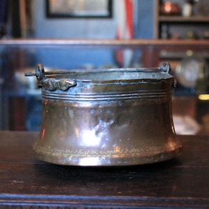 Może przedstawiać: Małe, antyczne wiadro miedziane z rozszerzoną podstawą i dwoma małymi uchwytami. Wiadro ma przyciemniony, postarzany wygląd, z widocznymi śladami zużycia i patyną. Obręcz posiada ozdobne poziome linie.