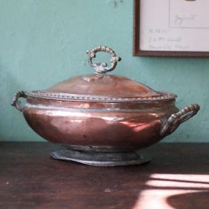Puede incluir: Una fuente de cobre con tapa y asas. La fuente tiene forma redonda con un borde decorativo y un pequeño asa en la parte superior de la tapa. La fuente se asienta sobre una base de color plateado. El fondo es de color verde azulado apagado.