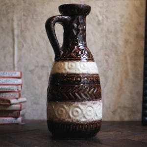 Puede incluir: Una jarra de cerámica alta con un esmalte marrón oscuro y dos bandas horizontales de patrones texturizados de color crema. La superficie presenta formas geométricas en relieve, incluyendo círculos, chevrones y diamantes, con un asa lateral.