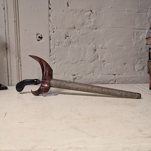 Puede incluir: Una daga de madera y metal con un mango curvo y una hoja plateada. La daga está apoyada sobre una superficie blanca.