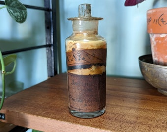 May include: A clear glass bottle with a brown label that reads "CALA" and "THE GREAT" in faded black lettering. The bottle is filled with a light brown substance and has a glass stopper.