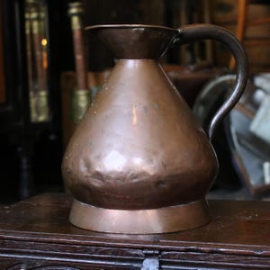 May include: A hammered copper pitcher with a wide, rounded body, a flared rim, and a curved handle. The metal surface displays a warm, metallic brown tone with subtle variations in texture and color across the exterior of the vessel.