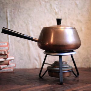 May include: A vintage copper fondue pot with a black handle and lid. The pot sits on a black metal stand with a burner below. The fondue pot is a warm copper colour.