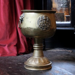 Può includere: Un grande vaso in metallo dorato con dettagli decorativi e uno stemma sul lato. Il vaso è su un piedistallo con una base rotonda.