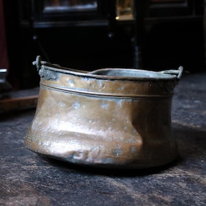 Peut inclure: Un seau en cuivre vintage avec une base légèrement évasée et deux petites poignées. Le métal vieilli a une couleur brun chaud avec des zones de vert-de-gris. Des accents de feuilles décoratives sont visibles près du bord.