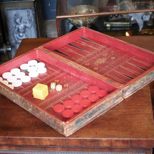 Puede incluir: Juego de backgammon abierto con interior rojo y exterior encuadernado en cuero. El juego incluye fichas blancas y rojas, dos dados y un dado amarillo. El juego se exhibe sobre una mesa de madera.