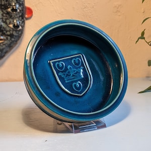 May include: A teal blue ceramic dish with a raised white crest featuring a crown and two hearts. The dish is on a clear acrylic stand.