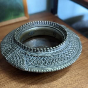 May include: A silver metal ashtray with intricate geometric designs. The ashtray has a round shape with a raised rim and a slightly indented center.