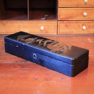 May include: A rectangular black box with a decorative lid featuring a gold and red Asian-inspired design. The box has a small circular clasp on the front and is set against a wooden surface. The box is likely used for storing small items.
