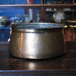 May include: A vintage brass bowl with a wide, rounded body and a slightly flared rim. The bowl has a warm, golden hue and shows signs of age and use. It sits on a dark wooden surface, suggesting a rustic or antique aesthetic.
