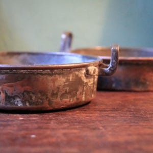 Könnte beinhalten: Zwei antike Kupferpfannen mit Griffen. Die Pfannen haben eine runde Form und weisen Gebrauchsspuren auf, mit einer rotbraunen Patina. Die gebogenen Griffe sind an den Seiten befestigt. Der Hintergrund ist verschwommen, mit einem Hauch von Grün.