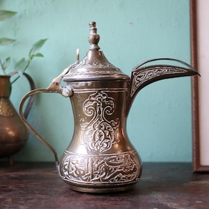 Puede incluir: Una cafetera antigua de latón con intrincados patrones grabados y un pico curvo. La cafetera presenta una tapa con un adorno decorativo y un asa para verter. El diseño incluye escritura árabe y motivos florales.