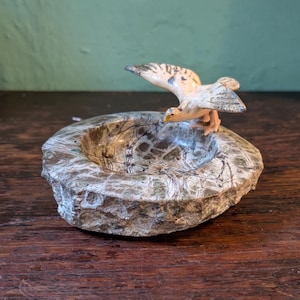 Puede incluir: Un cenicero decorativo con una base de piedra tallada y una figura de pájaro. El cenicero es redondo con una superficie texturizada y moteada en tonos grises, marrones y verdes. El pájaro es blanco con marcas grises y marrones.