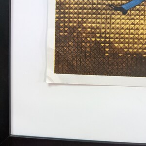 May include: A framed print with a black frame. The print features a geometric pattern in shades of brown and gold, with a blue element. The print is mounted on a white mat.