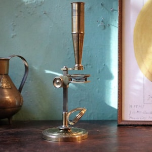 Könnte beinhalten: Ein antikes Messingmikroskop mit einem hohen zylindrischen Okular, einem zentralen Einstellknopf, einem flachen Objekttisch mit einem kleinen Federmechanismus und einem runden Fuß. Das Instrument hat ein goldenes Finish und eine vertikale Säule.