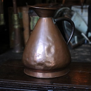 May include: A copper pitcher with a wide, flared rim and a dark metal handle. The body of the pitcher has a rounded, conical shape with a hammered, textured surface and a flat base. The metal shows a warm, reddish-brown metallic finish.