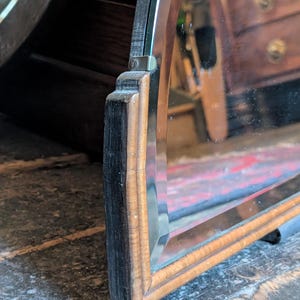 May include: A rectangular mirror featuring a beveled glass edge and a wooden frame. The frame consists of a dark, textured outer edge and a lighter, smooth inner border. A small metal bracket secures the glass to the wooden frame structure.