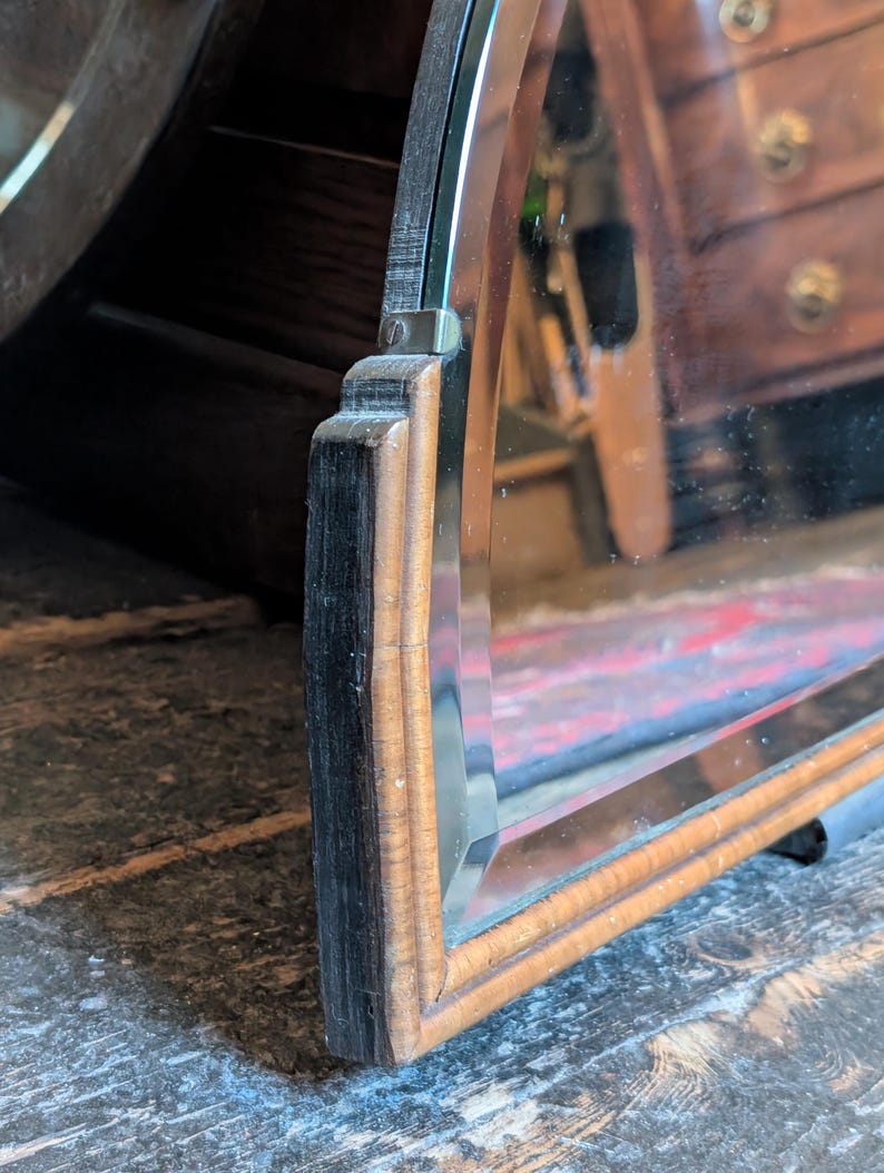 May include: A rectangular mirror featuring a beveled glass edge and a wooden frame. The frame consists of a dark, textured outer edge and a lighter, smooth inner border. A small metal bracket secures the glass to the wooden frame structure.