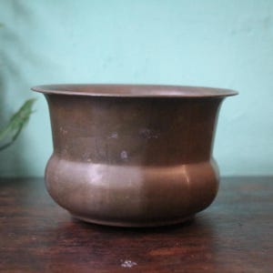 Peut inclure: Un petit pot de fleur antique de couleur bronze, avec une base arrondie et un bord évasé. Le pot est posé sur une surface en bois sombre, avec un fond vert clair. Le pot est vide et prêt à être planté.