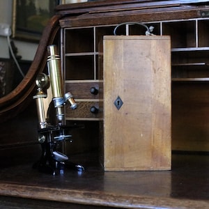 Puede incluir: Un microscopio de latón y metal negro con ocular y objetivo ajustables, situado junto a un estuche de almacenamiento de madera alto y rectangular con un asa de metal y una cerradura en forma de diamante en el panel frontal.