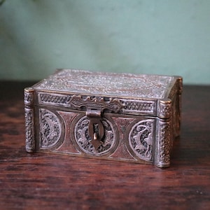 May include: A small, antique metal box with intricate embossed designs. The box features a hinged lid and a clasp closure. The metal has a tarnished, aged appearance, with detailed floral and geometric patterns. The box is rectangular and sits on a wooden surface.