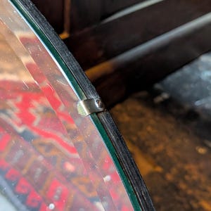 May include: A circular mirror featuring a dark wood frame with a beveled glass edge. A small, metallic silver-colored clip is secured to the frame with a screw, holding the glass pane firmly in place against the dark wooden border.