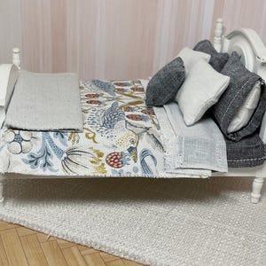 May include: Miniature white bed with a patterned quilt featuring birds and floral designs in blue, red, and gold. The bed is dressed with grey and white pillows and a grey blanket. The bed sits on a white rug.