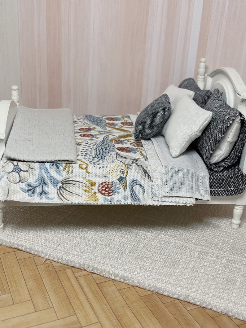 May include: Miniature white bed with a patterned quilt featuring birds and floral designs in blue, red, and gold. The bed is dressed with grey and white pillows and a grey blanket. The bed sits on a white rug.