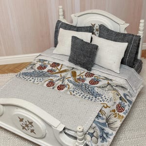 May include: Miniature white bed with a headboard and footboard. The bed is made up with a grey blanket and pillows. The blanket has a patterned design with birds and berries. The pillows are white and grey.