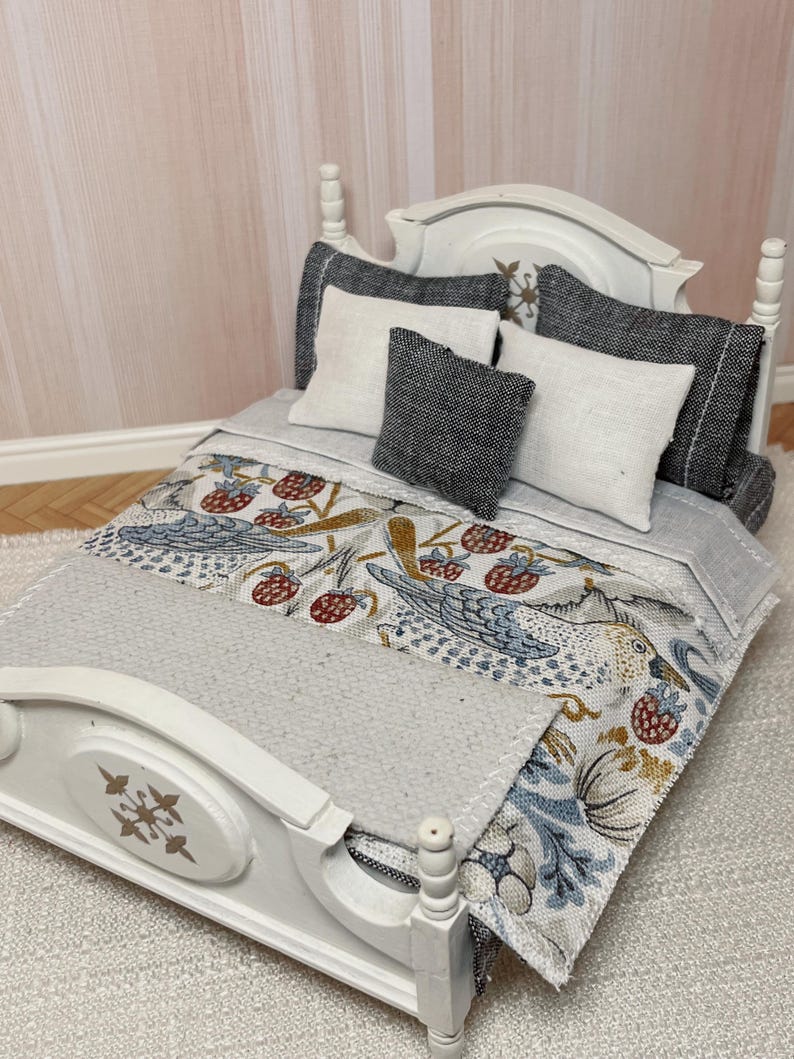 May include: Miniature white bed with a headboard and footboard. The bed is made up with a grey blanket and pillows. The blanket has a patterned design with birds and berries. The pillows are white and grey.
