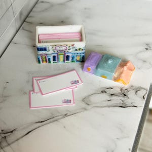 May include: A miniature stationery set featuring a small rectangular box with a colorful building design, filled with pink-edged cards. Several matching cards are scattered nearby, along with a small, colorful, house-shaped object.