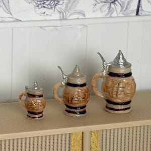 May include: Three miniature beer steins with silver lids and handles. The steins are brown with a textured design and black bands. They are arranged in a row, varying in size, and are set on a light-colored surface.