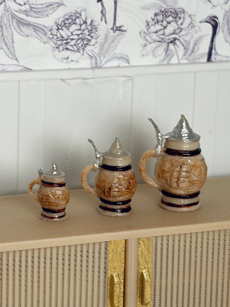 May include: Three miniature beer steins with silver lids and handles. The steins are brown with a textured design and black bands. They are arranged in a row, varying in size, and are set on a light-colored surface.