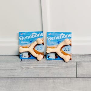 May include: Two Benebone wishbone dog chews on blue packaging. The chews are light tan and shaped like wishbones. The packaging has the Benebone logo and the words "wishbone" and "chicken". The background is a light grey wood.