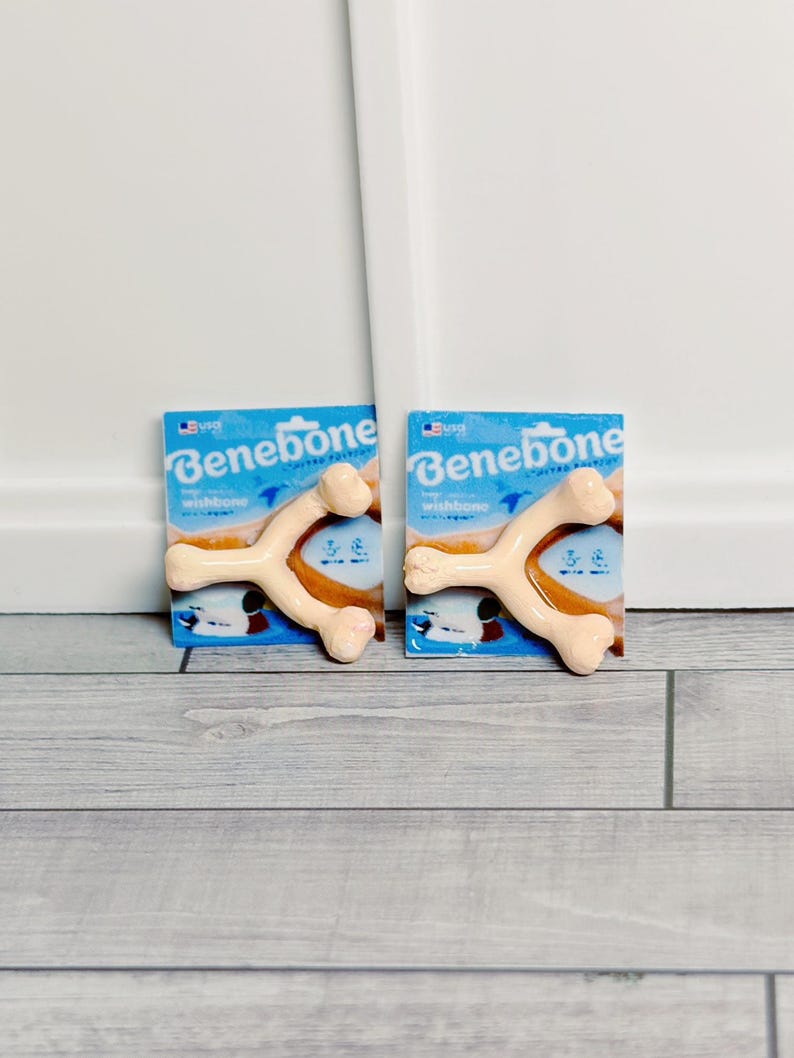 May include: Two Benebone wishbone dog chews on blue packaging. The chews are light tan and shaped like wishbones. The packaging has the Benebone logo and the words "wishbone" and "chicken". The background is a light grey wood.