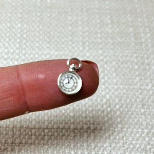 May include: A small, silver-colored pocket watch pendant with a white clock face and black Roman numerals. The watch has a loop at the top for hanging. The watch is held on a finger.
