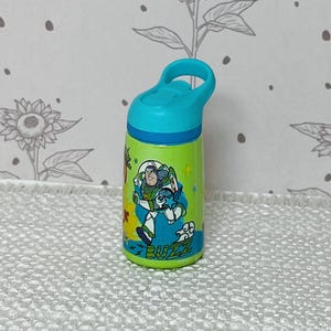 May include: A small, green water bottle with a blue lid and handle. The bottle features a colorful graphic of Buzz Lightyear from Toy Story, with the word "BUZZ" at the bottom. The background is a floral pattern.