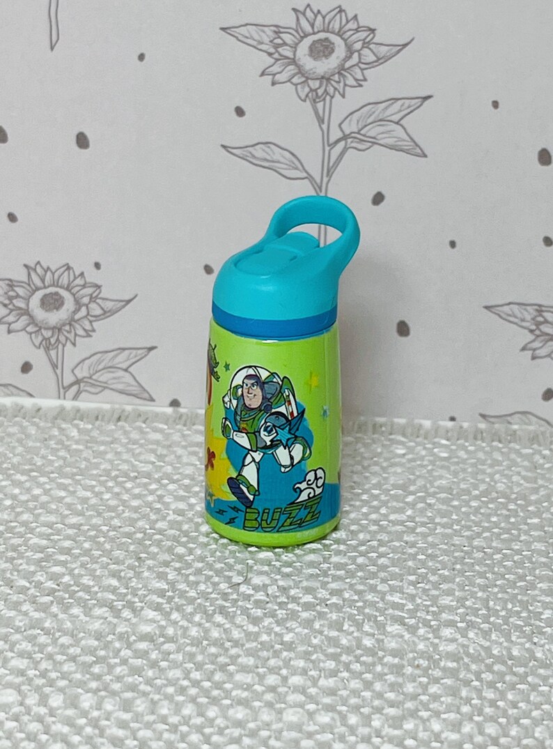 May include: A small, green water bottle with a blue lid and handle. The bottle features a colorful graphic of Buzz Lightyear from Toy Story, with the word "BUZZ" at the bottom. The background is a floral pattern.