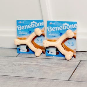 May include: Two Benebone wishbone dog chews in their packaging. The packaging is blue with white text and a graphic of the wishbone. The chews are tan and shaped like wishbones.