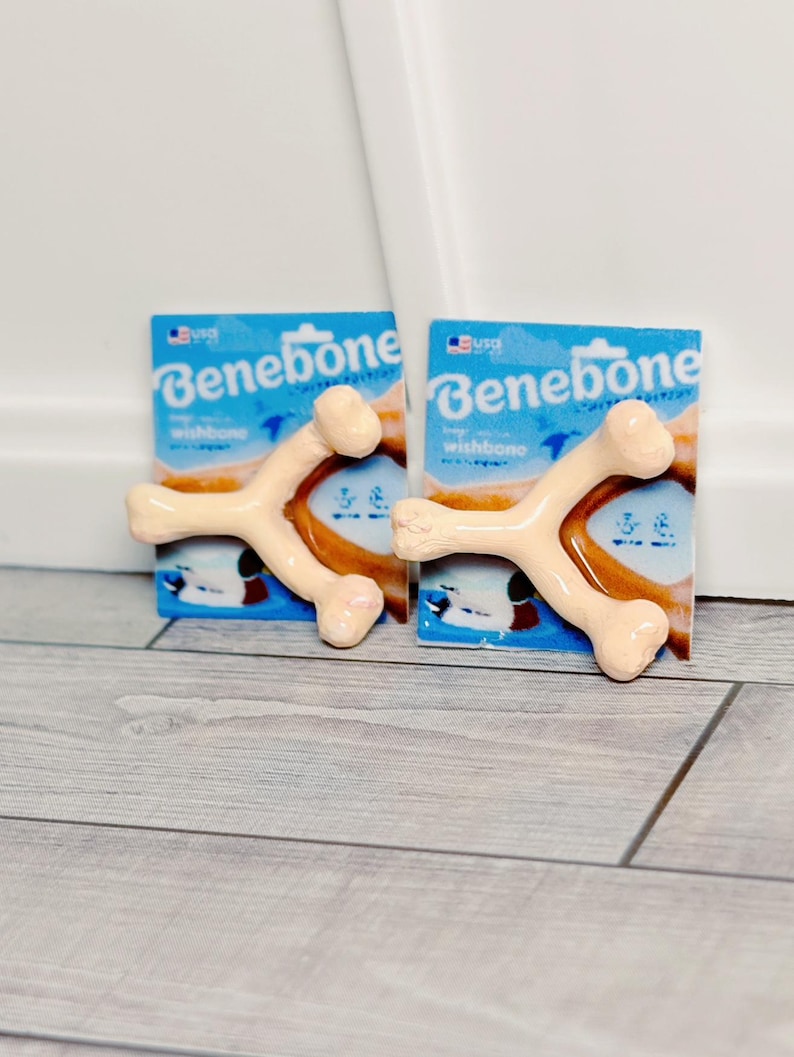 May include: Two Benebone wishbone dog chews in their packaging. The packaging is blue with white text and a graphic of the wishbone. The chews are tan and shaped like wishbones.