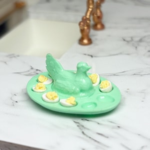 May include: A light green, oval-shaped serving dish with a duck figurine in the center. The dish holds several halved deviled eggs. The dish is on a white marble countertop, with a copper faucet in the background.